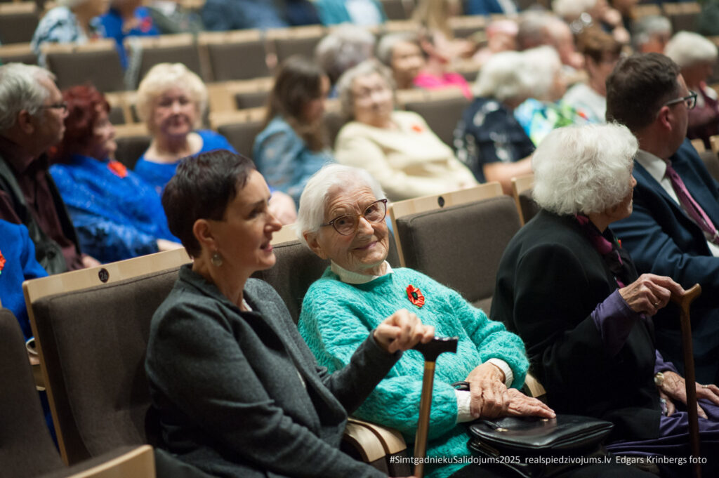 Foto no 2025. gada Simtgadnieku Salidojuma. Fokuss uz sievieti, kas skatās kamerā un smaida. Foto: Edgars Krinbergs